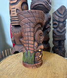 side view of a slipcase brown ceramic tiki mug with a detailed face, green leaves, and gold accents.