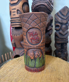 rear view of a slipcase brown ceramic tiki mug with a detailed face, green leaves, and gold accents.