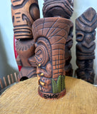 side view of a slipcase brown ceramic tiki mug with a detailed face, green leaves, and gold accents. slipcase brown ceramic tiki mug with a detailed face, green leaves, and gold accents.